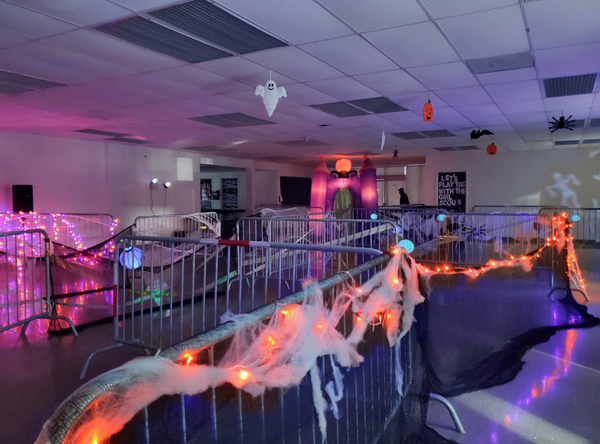 Ghosts, pumpkins, and spiders are hanging from the ceiling of a large room that is sectioned off with metal fencing that is wrapped in cob webs and lights. 