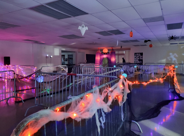 Picture2.jpg Ghosts, pumpkins, and spiders are hanging from the ceiling of a large room that is sectioned off with metal fencing that is wrapped in cob webs and lights.