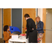 Two ladies stand and talk with a lady sitting at table by located by the door. 