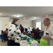 Lady in a sweater covered in hearts talking to a man holding a backpack. There are people on the other side of the room doing paperwork.