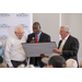  Three men stand with one speaking into a microphone reading from a framed certificate another man is holding.  